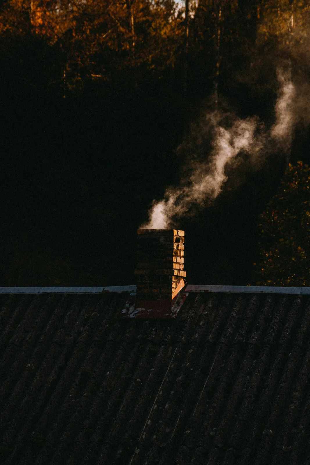 Smoke rising from a brick chimney on a shingle roof at dusk